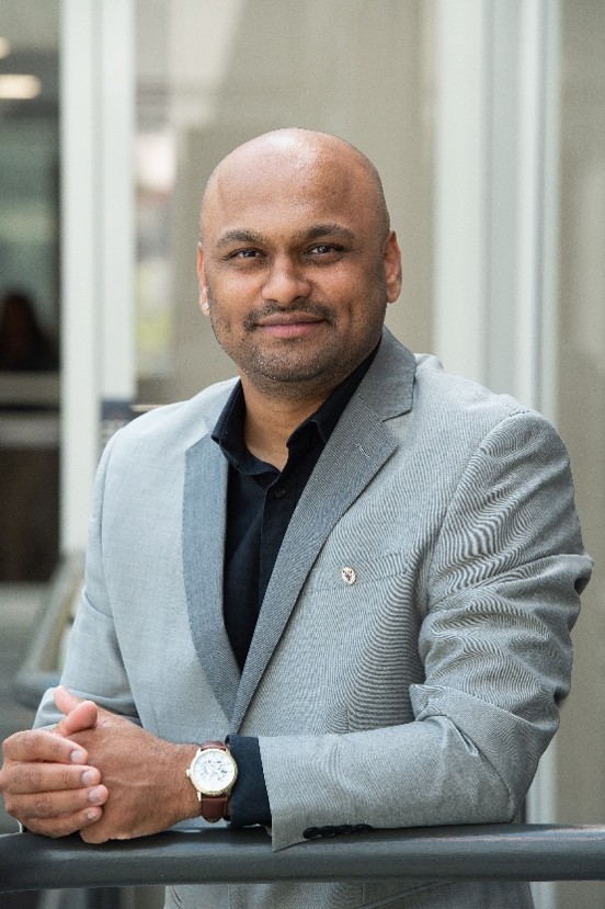 A professional image of a man in a grey suit jacket sitting down with his hands linked in front of him.