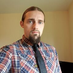 A picture of a man with long hair tied back neatly. He is wearing a checked shirt that is red, blue and white. He is wearing a black tie. He is sitting side on to the camera with his right arm closest to the camera.