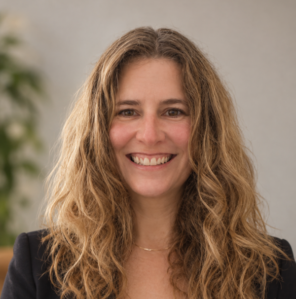 A smiling professional headshot of a woman with light wavy hair. There is a plant in the background.