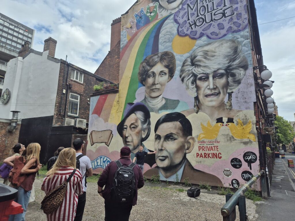 A painted mural on the side of a restaurant just off Canal Street in Manchester. The wall depicts several icons associated with LGBTQI+ in Manchester. The wall is colourful. A group of people are stood in front looking towards the mural.