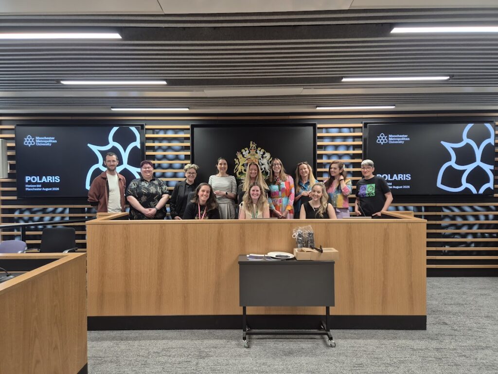 A group picture of several people behind a Judge's desk. The are screens behind to the left and right, and an image with a crest directly behind the people. They are smiling.