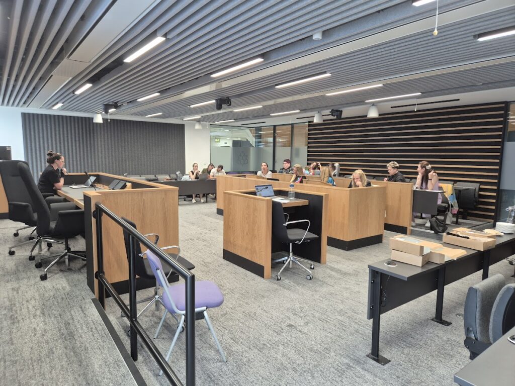 A different angle of the court room, from the right corner, with the judge bench to the left and the rest of the room directly in front. There are seats with tables and microphones to the front and sides of the bench, making it a square shape with the judges bench at the front. People are sat randomly around the room. One person is sat at the front. They look like they are having an active discussion.