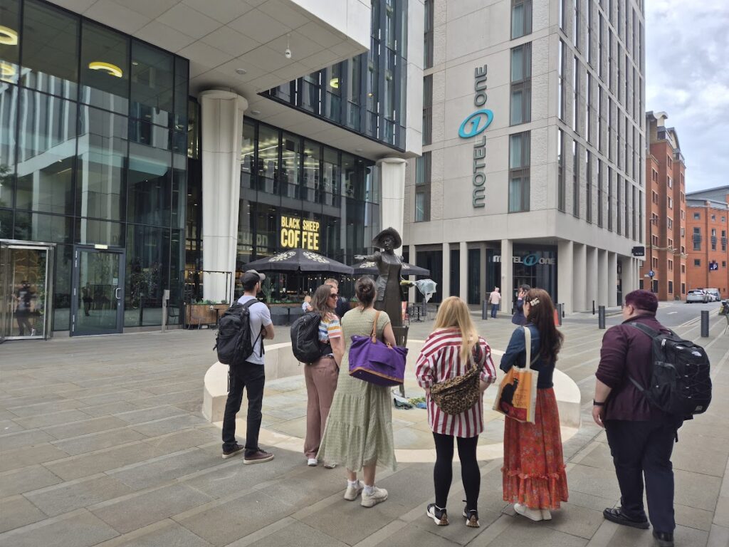 The scene is of a pedestrianised street with high rise buildings in central Manchester. The hotel in front says Motel One. There are a group of people in front of the Emmeline Pankhust statue. They are looking at the statue.